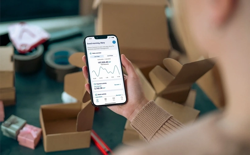 A person holding a smartphone displaying Elavon Connect with the greeting 'Good morning, Mary!' visible on the screen. In the background, there are cardboard boxes, tape, and small wrapped items, suggesting a packing or shipping environment.